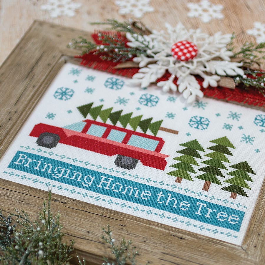 A photo of the Bringing Home The Tree Cross Stitch Pattern in a light wooden frame with holiday decor all around