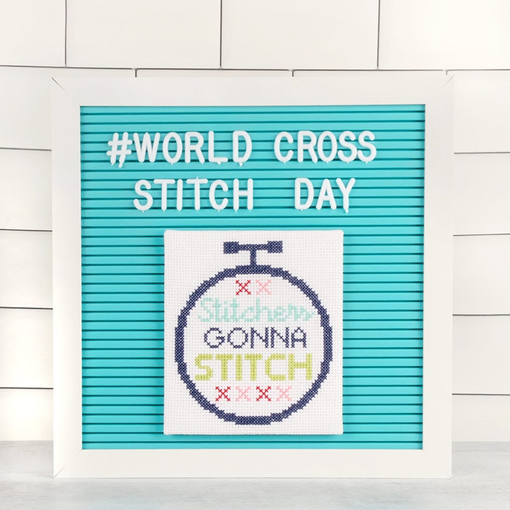 The Stitchers Gonna Stitch cross stitch pattern is finished on a blue announcement board, with white letters over head saying #World Cross Stitch Day 