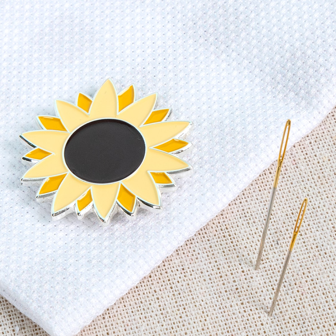 Image of a sunflower needle minder and a few cross stitch needles on a background of cross stitch cloth
